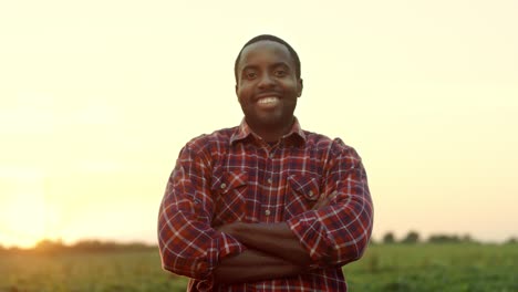 Retrato-Del-Apuesto-Joven-Agricultor-Afroamericano-Parado-En-El-Campo-Al-Atardecer,-Sonriendo-A-La-Cámara-Y-Cruzando-Las-Manos-Frente-A-él.